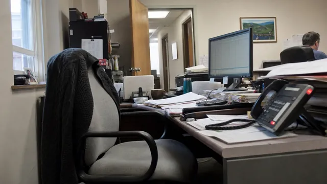 A cluttered small-office desk with an empty chair pushed back and a dark cardigan draped over it. In the foreground a desk phone has a red line indicator lit. Behind it are stacks of paper, a monitor showing a spreadsheet, and a coworker at another desk further back.