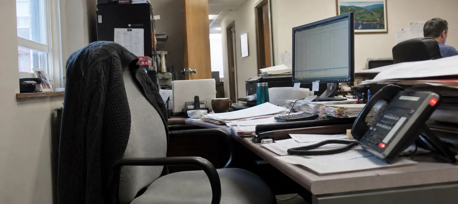 A cluttered small-office desk with an empty chair pushed back and a dark cardigan draped over it. In the foreground a desk phone has a red line indicator lit. Behind it are stacks of paper, a monitor showing a spreadsheet, and a coworker at another desk further back.