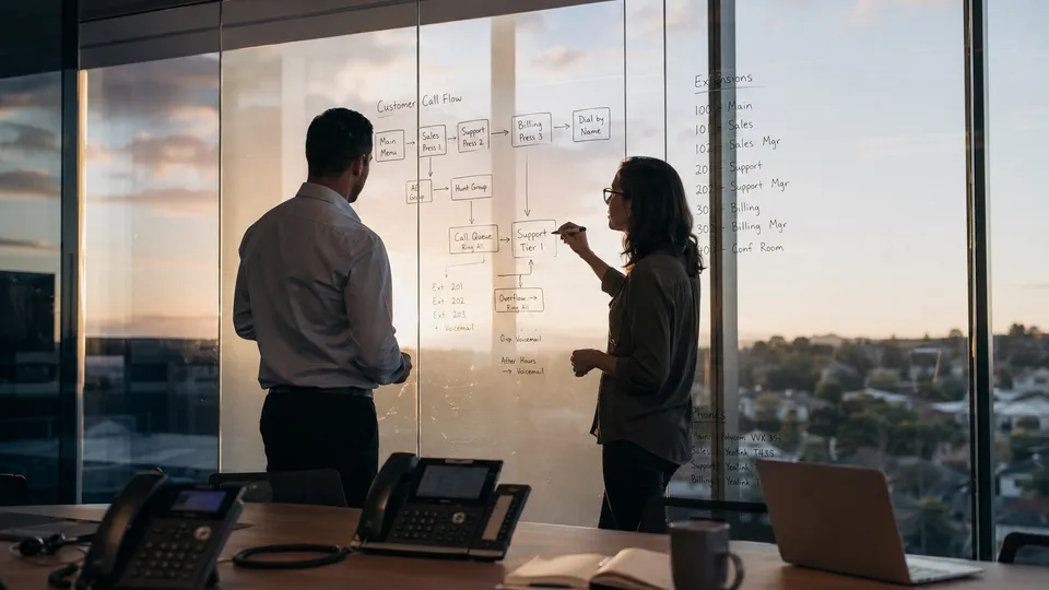 Two colleagues at a glass office wall mapping a customer call-flow diagram with extension assignments, business desk phones on the conference table in the foreground at dusk.